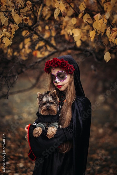 Fototapeta A young woman in a Day of the Dead costume: purple and violet skull makeup, red roses on a wreath, and a black cloak. She held a small dog against the backdrop of an autumn park with fallen leaves.