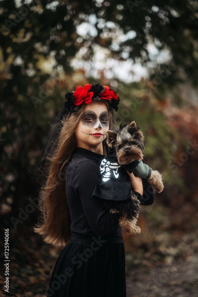 Fototapeta A young woman in a Day of the Dead costume: purple and violet skull makeup, red roses on a wreath, and a black cloak. She held a small dog against the backdrop of an autumn park with fallen leaves.