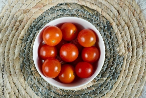 Obraz Cherry tomatoes fill a white bowl placed on a woven mat. Their vibrant red color contrasts with the neutral tones of the mat, highlighting their freshness and natural appeal.
