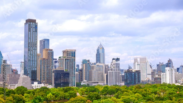 Fototapeta Modern city skyline and tall skyscrapers with green park on foreground against cloudy blue sky in Bangkok, Thailand. Cityscape view showing urban growth, architecture, and nature in harmony