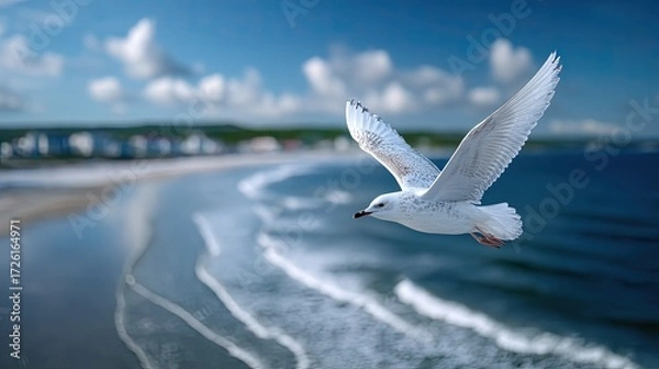 Fototapeta Seagull Mid Flight Above a Sandy Beach with Crashing Waves and Distant Buildings Under a Bright Blue Sky in a Cinematic HDR Wildlife Shot