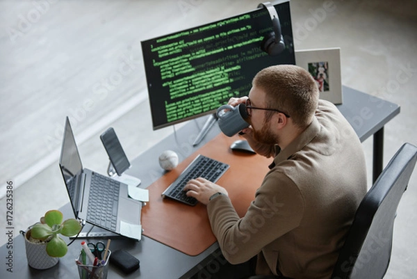 Fototapeta Caucasian young adult man working at desk using laptop and desktop computer with code on screen, holding mug and drinking while typing, sitting in modern office workspace