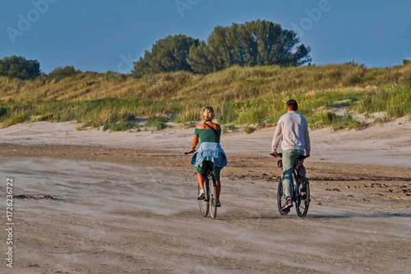 Fototapeta A couple riding bicycles on a sandy beach near grassy dunes during a sunny day. Active lifestyle and leisure at the seaside.