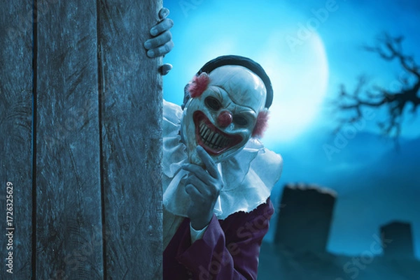 Fototapeta Man wearing scary clown mask peeking behind wooden wall on graveyard at night with full moon, Haloween theme