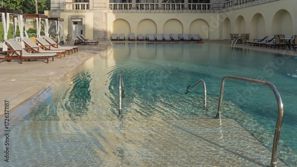 Fototapeta Swimming resort pool. Clear water, turquoise tiled bottom, metal handrails of stairs. Rows of deck chairs and awnings with curtains around the perimeter. Cuba. Havana. Hotel