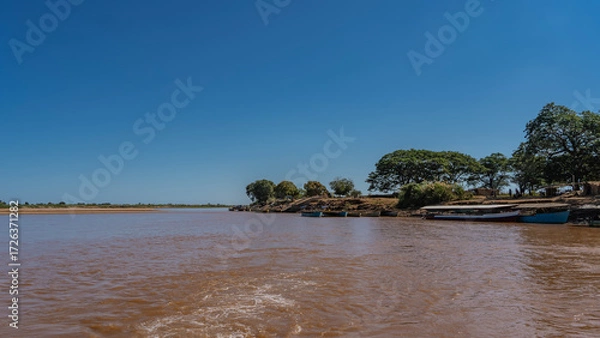 Fototapeta An exotic African river. A foam trail on brown water. The boats are moored at the shore. Green trees on the coast. Clear blue sky. Copy space. Madagascar. Tsiribihina river.