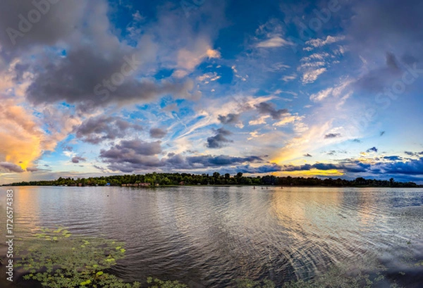 Fototapeta An evening sunset of intermingled cloud patches creates a layered, three-dimensional panoramic picture. The interplay of orange and blue tones. The river reflects the colors of the sky in its waves.