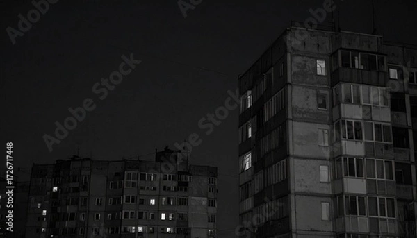 Fototapeta Apartment buildings stand against a dark night sky, with some windows illuminated, conveying a sense of urban living, isolation, and a hint of activity in the darkness.
