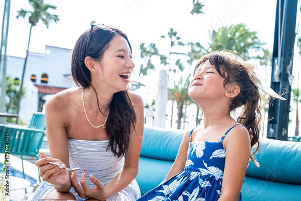 Fototapeta Japanese mother and daughter laughing together at a resort poolside café during summer vacation with palm trees bright sunlight and joyful tropical family travel atmosphere