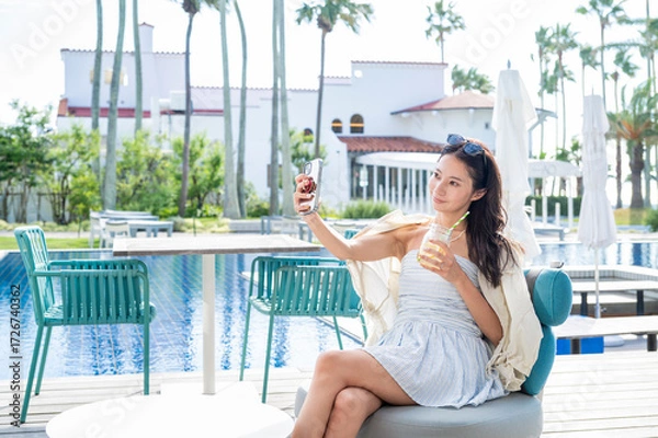 Fototapeta A Japanese woman sitting by a resort swimming pool holding a cold drink and taking a selfie with her smartphone while enjoying a relaxing summer holiday