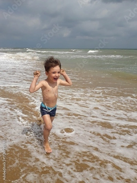 Fototapeta Young caucasian child joyfully running on sandy beach with waves in background