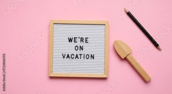 Fototapeta A small, light-wooden letter board displays "We're On Vacation". A small wooden tool and pencil rest beside it on a pink surface