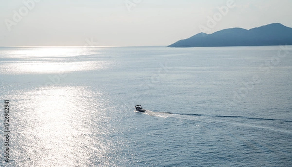 Fototapeta a vast expanse of shimmering sea surrounded by a chain of mountains under a partly cloudy sky. A boat is cutting through the water, leaving a trail behind. The sunlight filters through the clouds