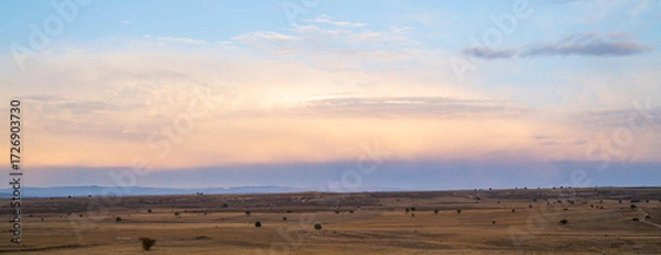 Fototapeta Expansive view of a harvested field at sunset with scattered trees and distant mountains, under a vast sky showing signs of rain in a rural landscape during early autumn