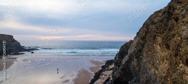 Fototapeta A peaceful coastal scene at sunset with a solitary figure walking on the sandy beach. The waves gently lap the shore, and towering cliffs frame the coast of Portugal.