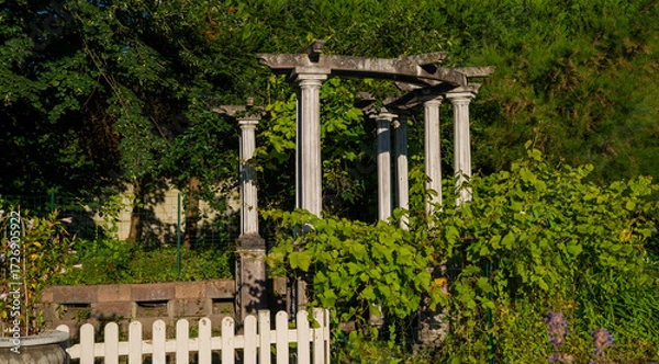 Fototapeta Ancient stone columns entwined with lush green vines stand in a peaceful garden, bathed in soft sunlight. The serene scene is framed by greenery, the classical architecture and natural beauty.