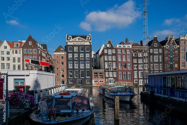 Fototapeta Narrow canal houses with gabled roofs and large windows near a canal in a sunny urban setting, featuring glass-roofed boats docked in the foreground and a bright blue sky above.