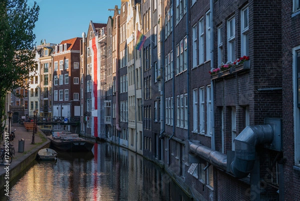 Fototapeta Narrow canal lined with tall brick houses in the historic center of Amsterdam, Netherlands. A moored boat, bicycles, and rainbow flag add life to the scene in soft evening light.