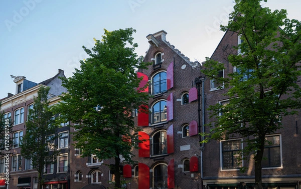 Fototapeta Red shutters accent the arched windows of a historic brick warehouse in central Amsterdam, Netherlands. The gabled roof lines and leafy green trees frame this iconic canal district architecture