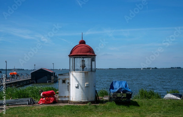 Fototapeta Small vintage lighthouse with a red dome and white base stands on a grassy shore by the Markermeer lake in Marken, North Holland. Sailboats, a pier, and boat storage structures are visible nearby.