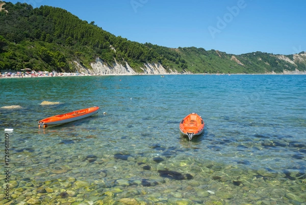 Fototapeta Sunny day at the beach across the cliffs in Portonovo Beach, Marche, Italy. Two red boats on the waves across the cliffs and sea