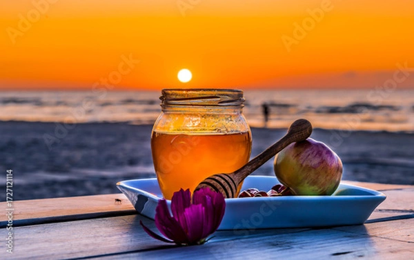 Obraz Close up with jar of honey and apples  - traditional food for Jewish New Year holiday - Rosh Hashanah. Focus on food jar with honey, blurred sand beach with people silhouette, sea waves and sunset