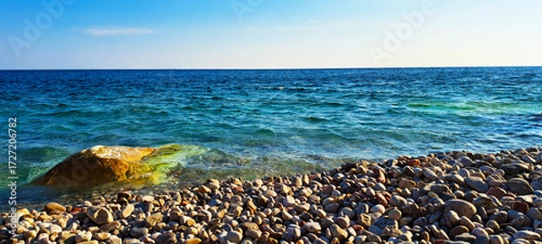 Obraz Scenic view of a rocky seashore with clear turquoise water, sunlit waves, and submerged stones creating a vibrant coastal landscape under a bright sky.