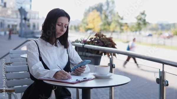Fototapeta A business woman is seated at a cafe table, using her phone and taking notes. She appears focused on her work while enjoying a coffee in a lively outdoor setting with people passing by