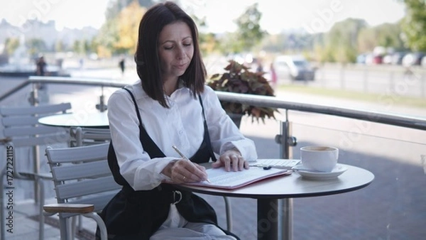 Fototapeta A woman sits at a table outside, focused on writing in her notebook. She enjoys a cup of coffee while surrounded by a lively atmosphere on a sunny day in a bustling area