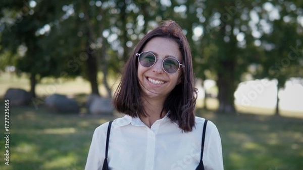 Fototapeta A confident businesswoman stands outdoors, smiling brightly. She has dark hair and sunglasses on, enjoying a sunny day in a park-like environment with trees and rocks nearby