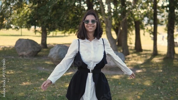 Fototapeta A woman dressed in a stylish outfit stands outdoors, smiling broadly with arms outstretched. She enjoys the warm sunshine and fresh air in a natural setting with trees and rocks