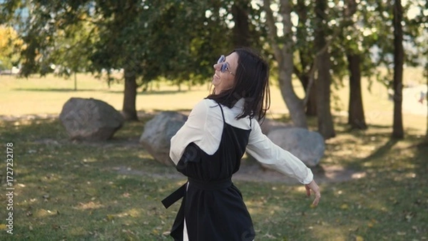 Fototapeta A confident woman in business attire stands in a park, smiling as she interacts with her surroundings. The bright sunlight adds warmth to the scene, enhancing the relaxed atmosphere of the moment