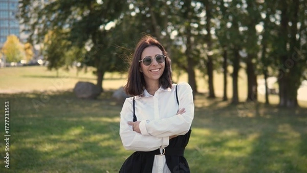 Fototapeta A woman stands confidently in a green park, wearing sunglasses and a stylish outfit. The sun shines down as she smiles, enjoying a warm day among trees and grass
