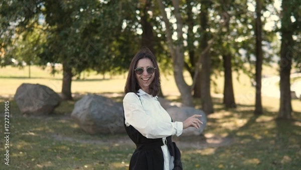 Fototapeta A cheerful businesswoman stands in a park surrounded by trees and rocks. She smiles confidently while wearing stylish sunglasses and a fashionable outfit on a bright day