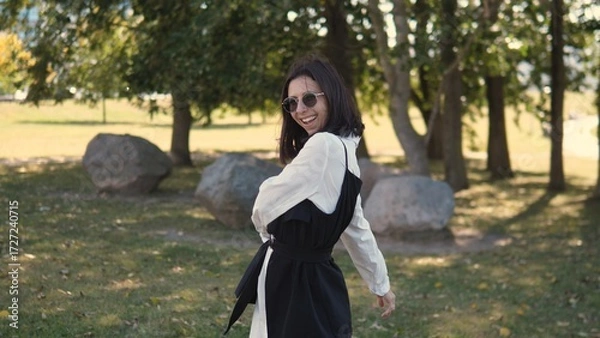 Fototapeta A businesswoman stands outdoors in a park, wearing stylish sunglasses and a casual outfit. She smiles brightly while surrounded by greenery and large rocks, enjoying a sunny day