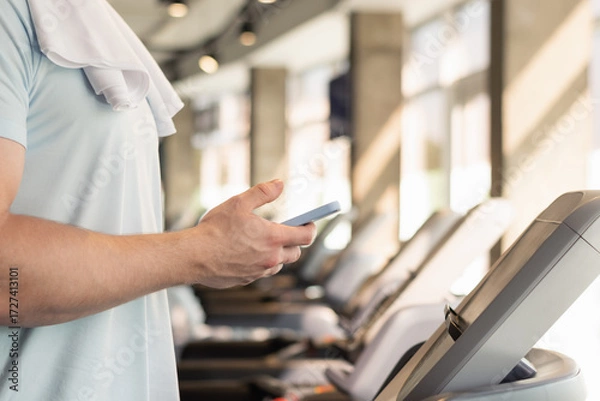 Fototapeta Hands sporty fit man with mobile phone checking apps and fitness progress on running track. Young athlete warm up on treadmill at gym. Close up.