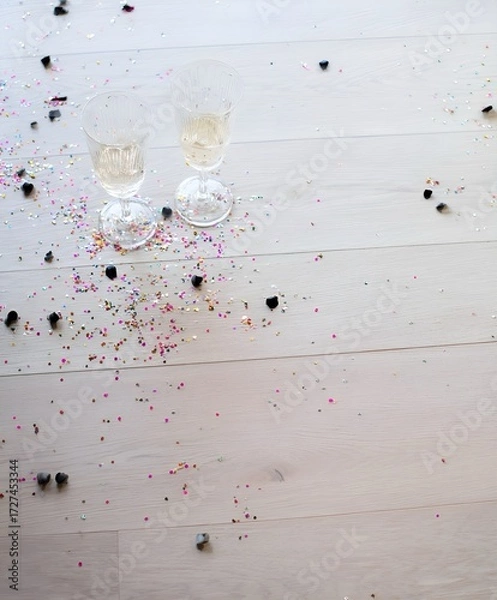Fototapeta Two elegant champagne glasses standing on a light wooden floor surrounded by colorful confetti and black decorative pieces after a celebration. Bright soft daylight and festive mood.