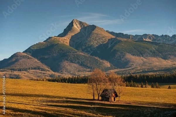 Fototapeta A small wooden cabin in a golden autumn meadow beneath a towering mountain peak, bathed in warm evening sunlight in a scenic wilderness landscape.