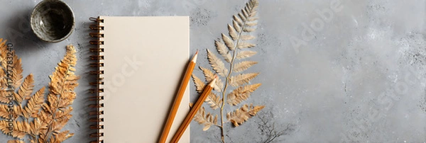 Fototapeta Flat lay of a spiral-bound notebook and two pencils next to dried brown ferns, resting on a textured gray concrete background, symbolizing writing, nature, and creative planning.
