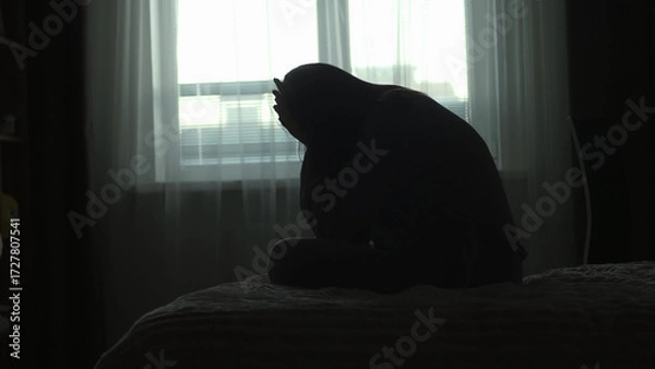 Obraz Silhouette of a millennial woman sitting on a bed in the dark bedroom, depression, pain. Portrait of Unhappy sad woman sitting against window and thinking about her life