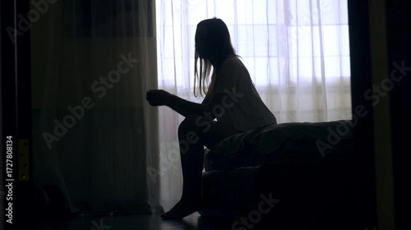 Obraz Silhouette of unrecognized woman sitting on a bed in the dark bedroom, depression, pain. Portrait of Unhappy sad woman sitting against window and thinking about her life