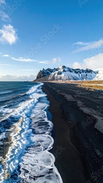Obraz Aerial view of a black sand beach meeting a wave-lapped ocean under a clear blue sky, snow-capped mountains in the background