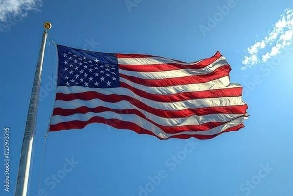 Obraz American flag waving strongly on a tall flagpole against a clear blue sky with a few scattered clouds