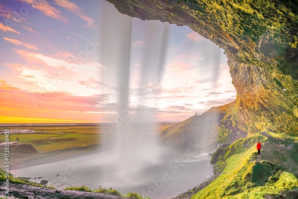 Fototapeta Sunset behind Seljalandsfoss waterfall, Iceland, showing cascading water, colorful clouds, and person in red admiring the dramatic landscape from the trail below.