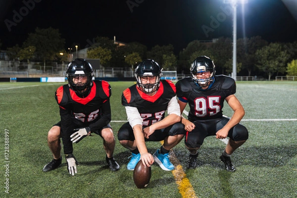 Fototapeta American football players in uniform and helmets crouching on a green turf field at night, ready for competition