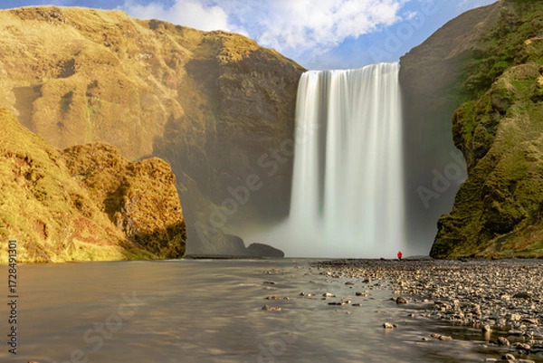 Fototapeta Long exposure view of Skógafoss waterfall, Iceland, flowing into a rocky river foreground. A lone traveler in red jacket adds dramatic sense of adventure and scale.