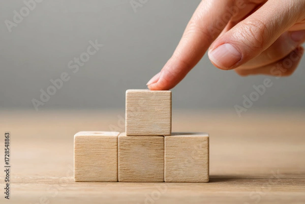 Fototapeta Hand Placing Wooden Block on Stack of Blocks on Wooden Surface with Gray Background