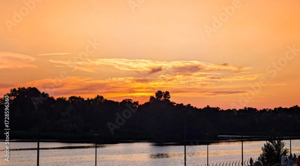 Fototapeta A beautiful yellow sunset over a calm river. Several clouds frame the setting sun. A row of trees outlines the forest edge on the horizon. Beautiful colorful dramatic sky with cloud.