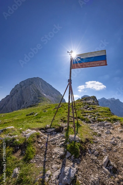 Fototapeta Scenic view from the  sun backlit Slovene flag on Kamniško sedlo in the Kamnik-Savinja Alps, Slovenia, showcasing alpine peaks like Brana and Turska gora, summer landscape