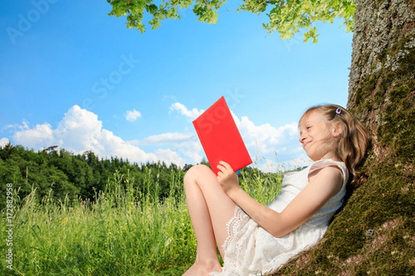 Obraz young girl sit next to a tree and smile into the summer sun. she plays with electronic devices, red apple, flowers. sky is blue and the grass is green.